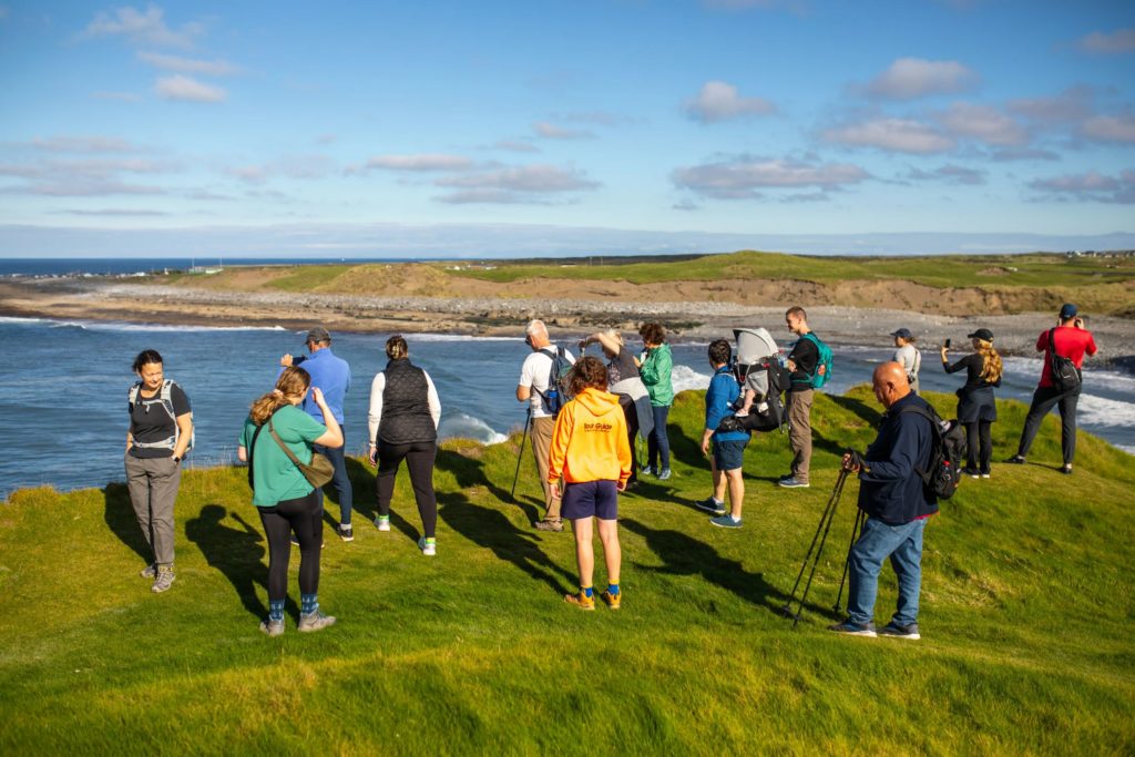 AKphoto (018) People enjoying scenic coastal view, capturing photos on a grassy cliff under a clear blue sky.