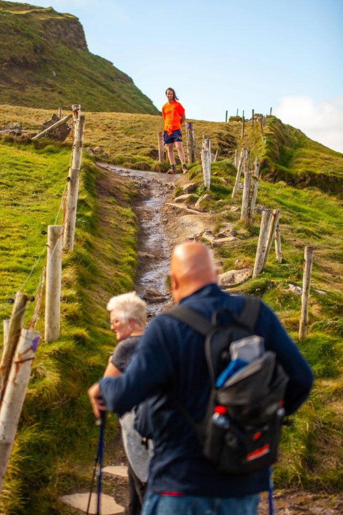 AKphoto (102) Hikers on a scenic, grassy trail enjoying a sunny day in the hills, with one person in a bright orange shirt ahead.