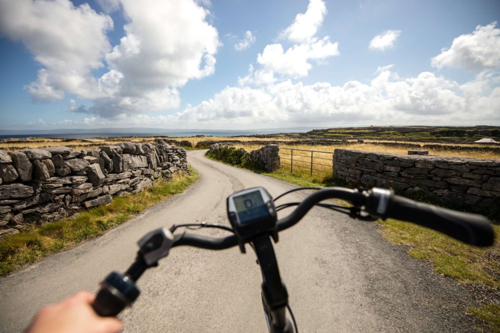 AKphoto (116) Cyclist rides on a scenic rural road in Ireland, flanked by stone walls and fields under a blue sky with clouds.