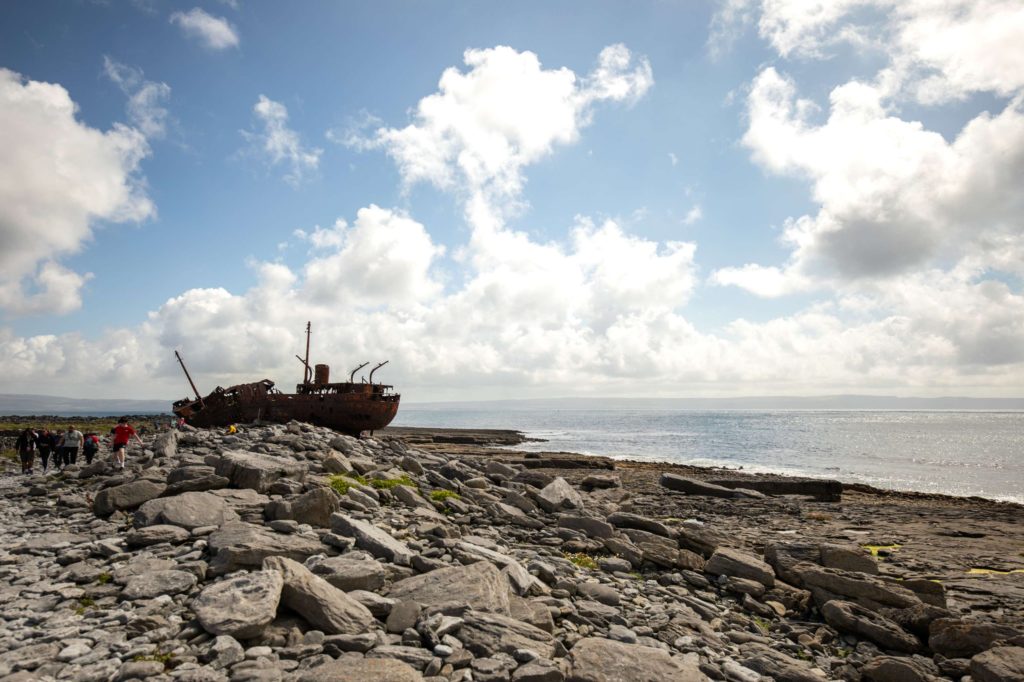AKphoto (119) Rusty shipwreck on rocky shore under bright sky, ocean in the background, and a group of tourists exploring the landscape.