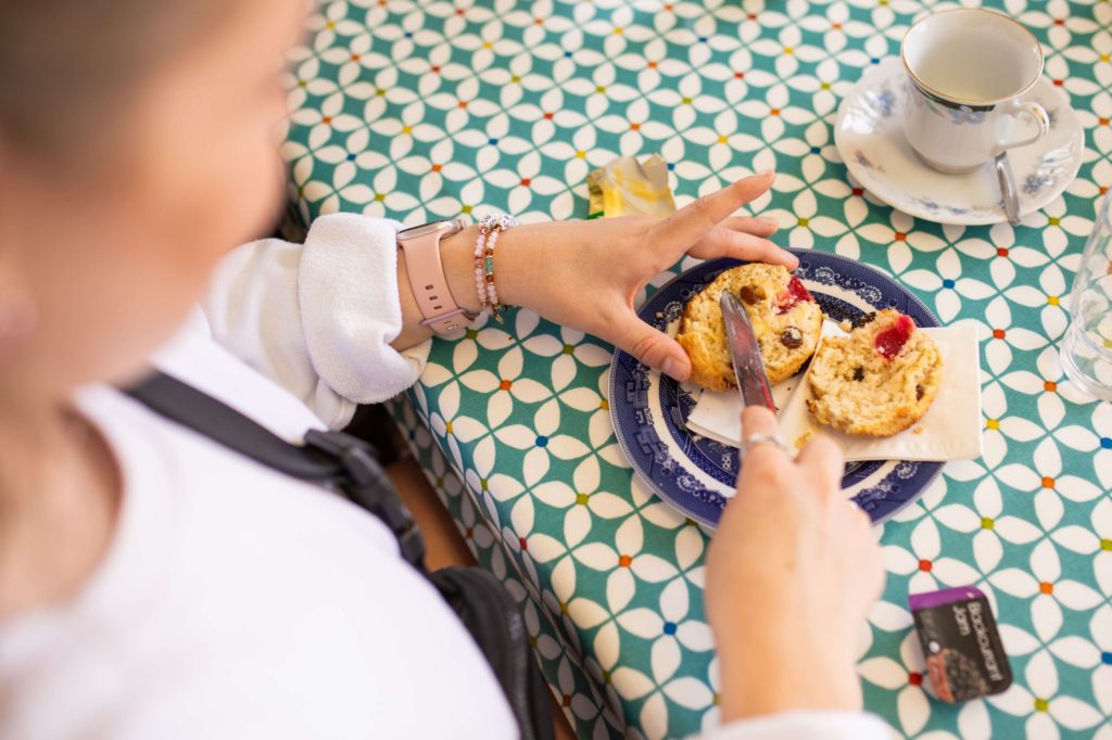 AKphoto (12) Person spreading jam on a scone with a cup of tea nearby on a patterned tablecloth.