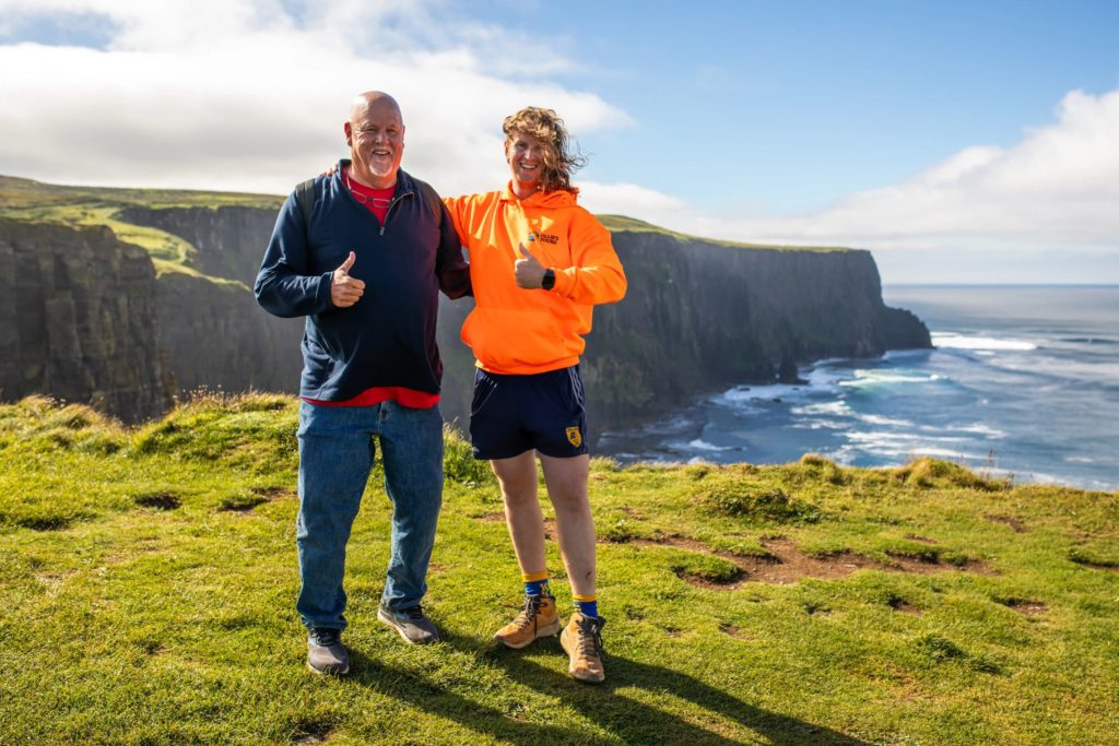 AKphoto (127) Ollie and tourist smiling with thumbs up at the Cliffs of Moher, Ireland, on a sunny day with a coastal view.