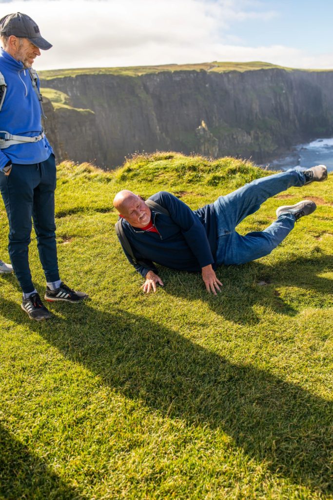 AKphoto (129) Man jokingly posing on grass near coastal cliffs on a sunny day, while another man stands beside him smiling.