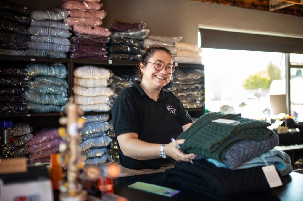 AKphoto (155) Shop assistant smiling while arranging knitted jumpers in a yarn shop, with shelves of colourful wool behind her.