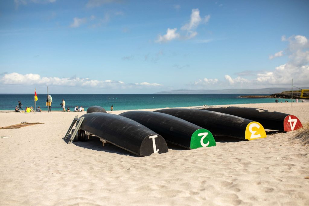 AKphoto (159) Four upside-down canoes with colourful ends on a sandy beach near a calm sea under a blue sky.