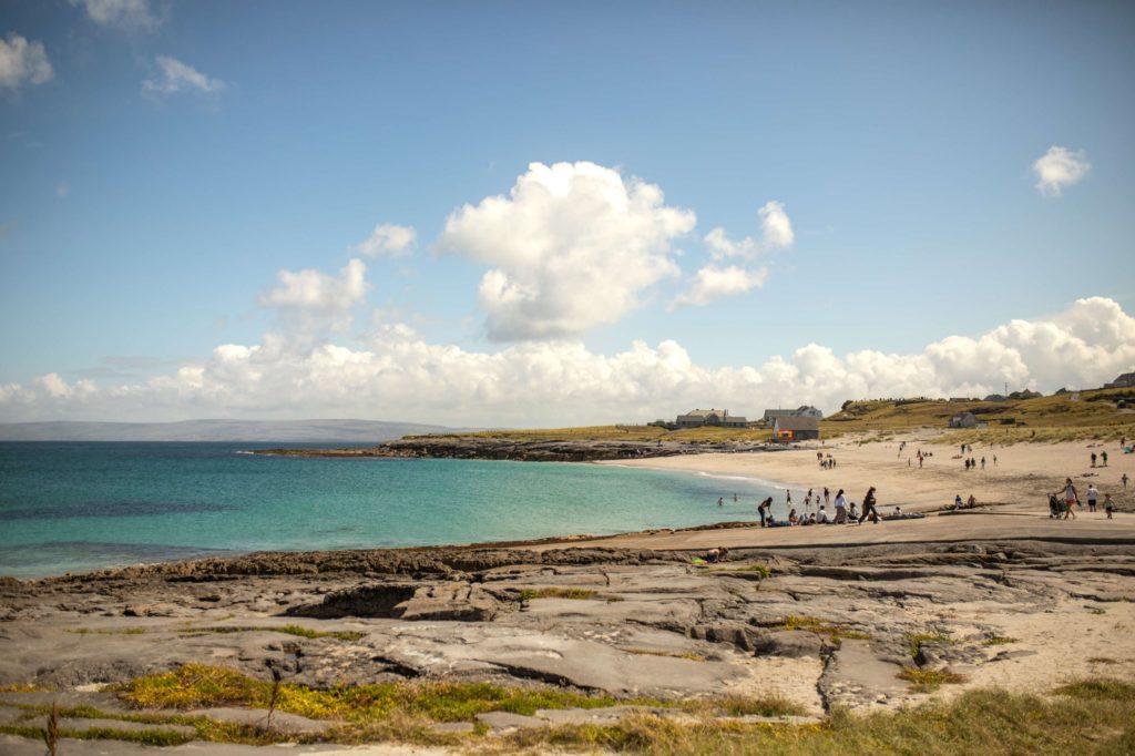 AKphoto (161) Scenic beach with turquoise sea and cloudy sky, rocky foreground, and people enjoying a sunny day near distant buildings.
