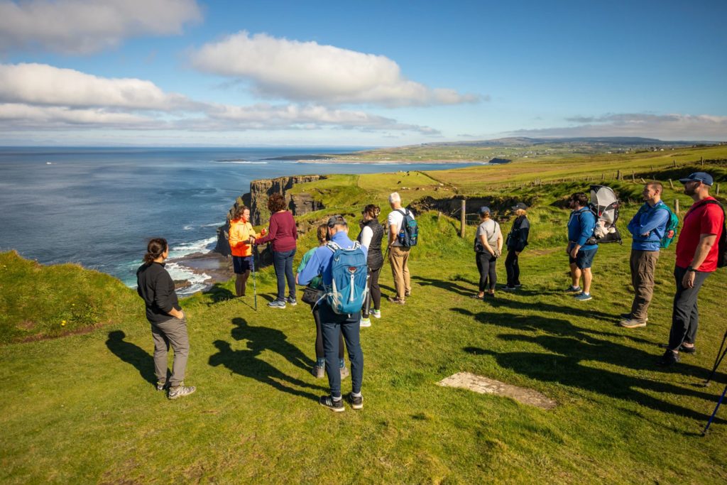 AKphoto (167) Group of hikers enjoying a scenic view on a grassy cliffside overlooking the ocean under a blue sky.