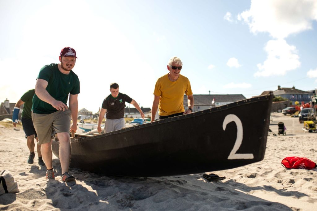 AKphoto (170) Men pulling a wooden boat onto a sandy beach on a sunny day, with buildings in the background.