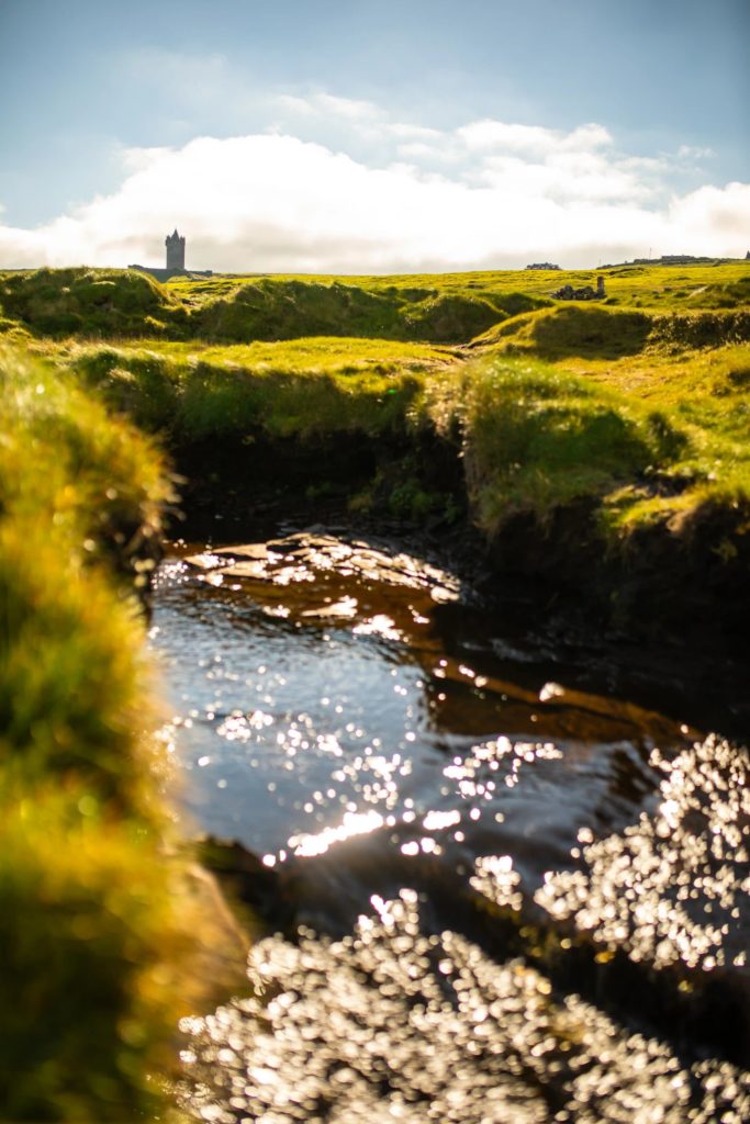 AKphoto (33) Sunny Irish landscape with glistening stream and distant tower against a bright sky.