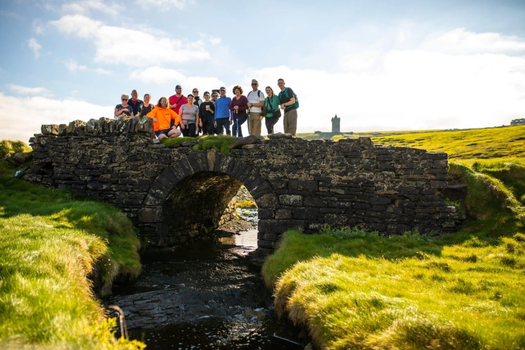 AKphoto (41) Group of people on a historic stone bridge in a scenic, grassy landscape with an ancient castle in the distance.