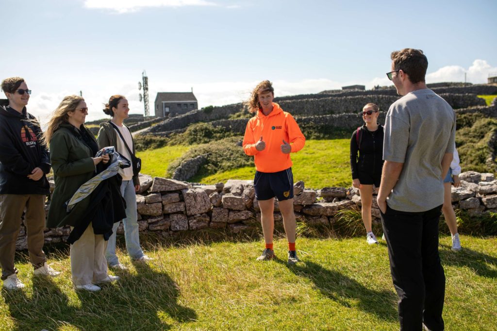 AKphoto (48) Group of people enjoying a sunny countryside tour, gathered near a stone wall with a guide in an orange hoodie.
