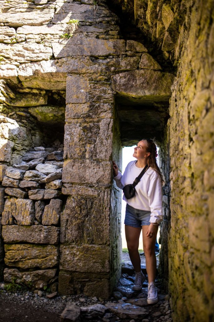 AKphoto (65) Young woman exploring ancient stone ruins in casual outfit, looking upwards with curiosity and wonder.