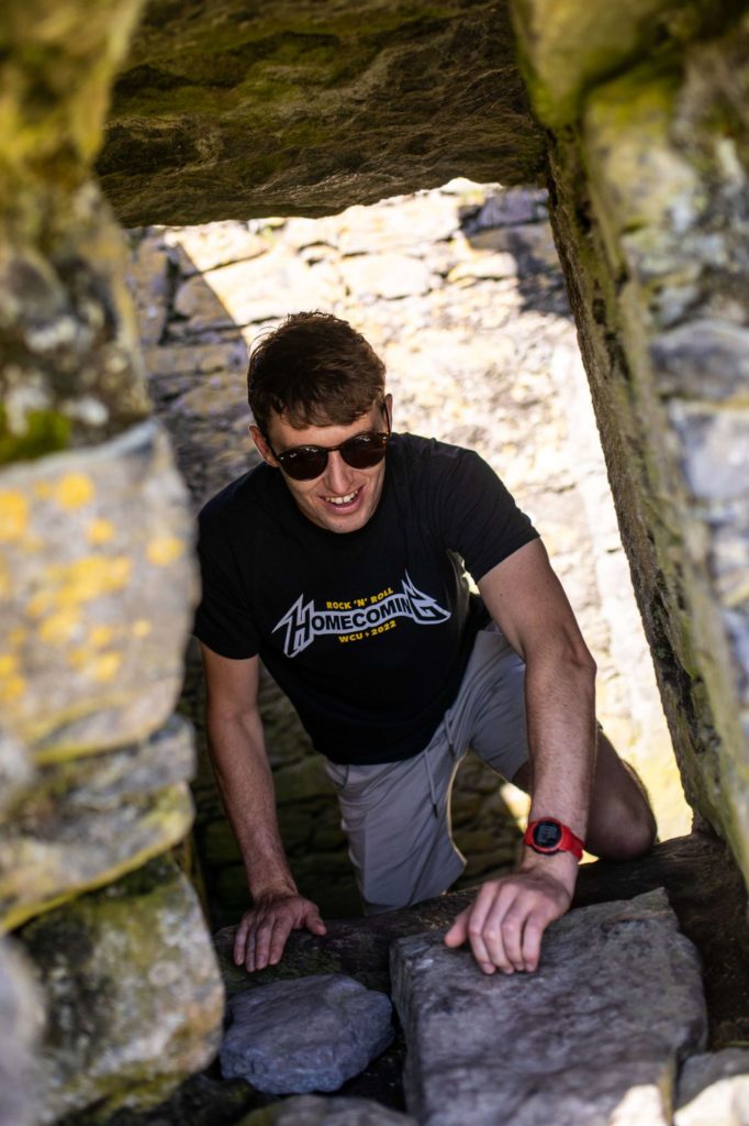 AKphoto (68) Man wearing sunglasses explores a stone ruin passageway, smiling in the sunlight. Adventure and history blend outdoors.