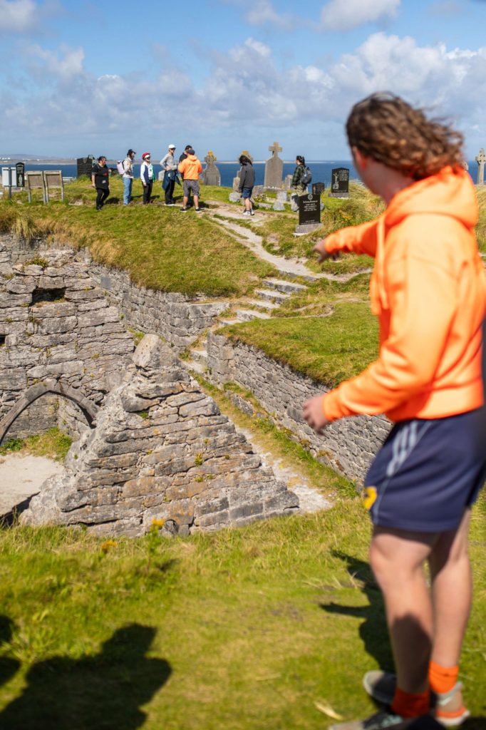 AKphoto (78) Ollie points at a group of tourists on grassy hill near ancient ruins and gravestones with coastal view.