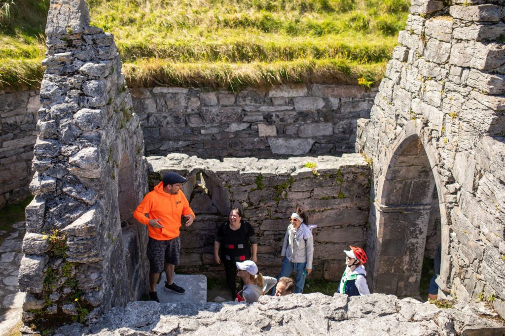 AKphoto (80) Tour group exploring ancient stone ruins under a sunny blue sky, led by a guide in a bright orange hoodie.