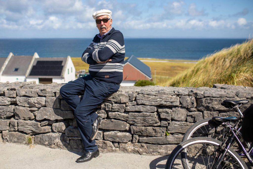 AKphoto (88) Elderly man in cap and sunglasses leans on stone wall by the sea, with bicycle and cottages in background.