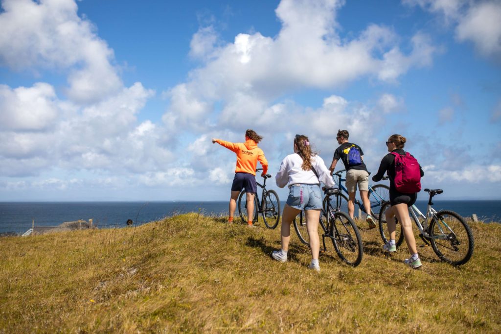 AKphoto (89) Group of cyclists on a scenic coastal path under a clear blue sky, overlooking the sea, enjoying an outdoor adventure.